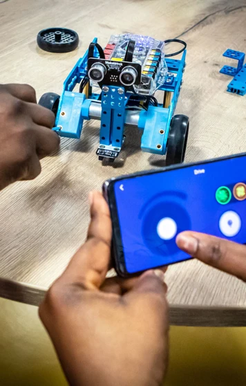 Student using a smartphone to control a small blue robotic vehicle, testing its movements on a table in a computing lab. Student using a smartphone to control a small blue robotic vehicle, testing its movements on a table in a computing lab.